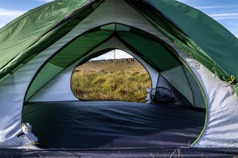 Axis-2-1-3.jpg Interior view of a green-and-white dome tent with the entrance unzipped, the opening framing a grassy meadow and low rocky ridge under a blue sky.