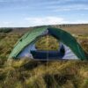 Green dome tent with its entrance open, staked and yellow guy‑lined on windswept grassy moorland with a low rocky escarpment and blue sky in the background.