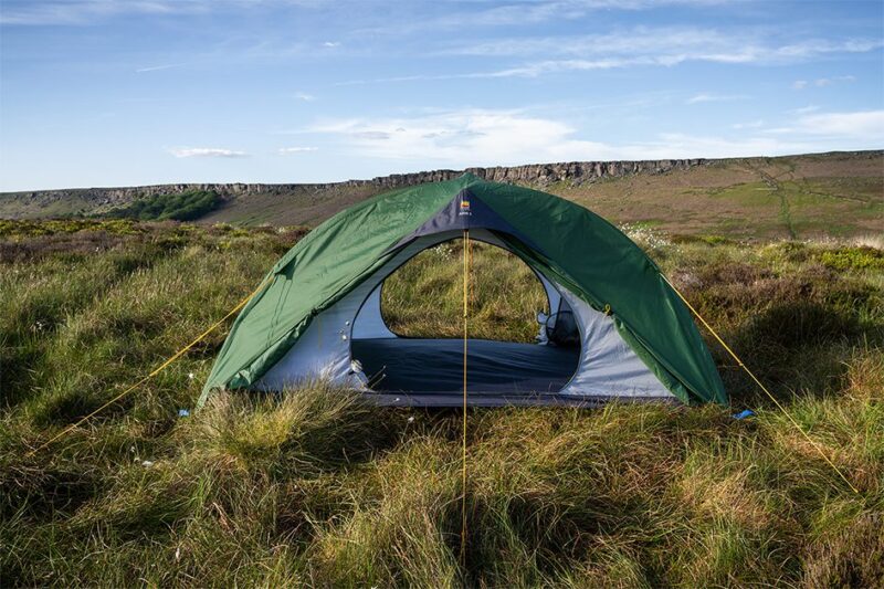 Axis-2-2-1-3.jpg Green dome tent with its entrance open, staked and yellow guy‑lined on windswept grassy moorland with a low rocky escarpment and blue sky in the background.