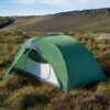 Green two-person dome tent with its front mesh door open, pegged on grassy moorland among low heather and white cotton‑grass, with a low rocky escarpment and blue sky in the background.