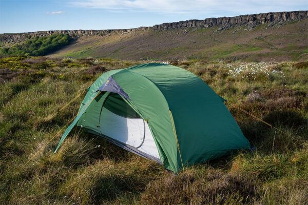 Green two-person dome tent with its front mesh door open, pegged on grassy moorland among low heather and white cotton‑grass, with a low rocky escarpment and blue sky in the background.