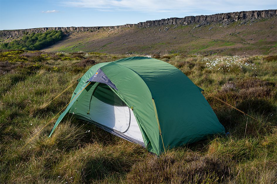 Green two-person dome tent with its front mesh door open, pegged on grassy moorland among low heather and white cotton‑grass, with a low rocky escarpment and blue sky in the background.