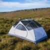 Pale blue-and-white dome tent with mesh roof and exposed poles pitched on grassy moorland, with a low rocky escarpment and blue sky in the background.