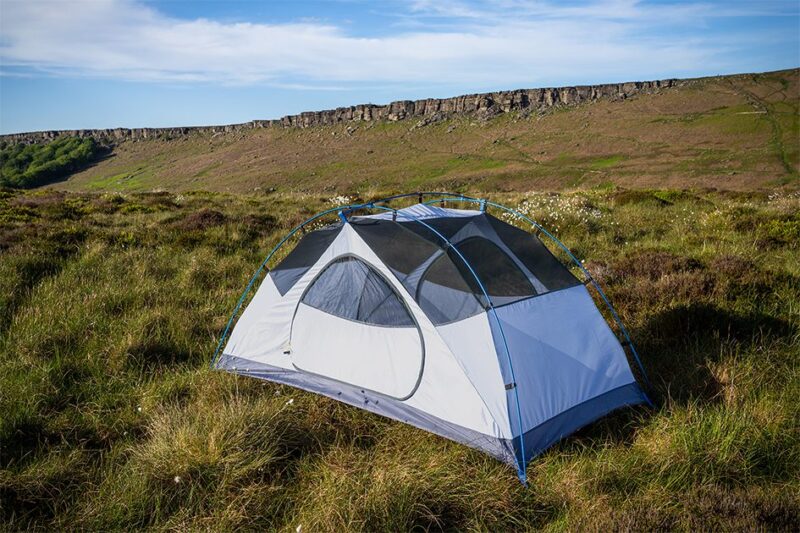 Axis-2-4-3.jpg Pale blue-and-white dome tent with mesh roof and exposed poles pitched on grassy moorland, with a low rocky escarpment and blue sky in the background.