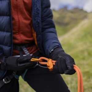 Close-up of a person in a navy puffer jacket and black gloves threading an orange climbing rope through an orange belay device and carabiner while clipped into a harness on a grassy hillside