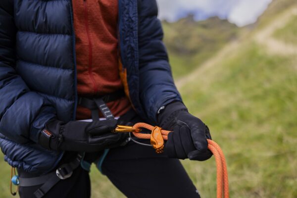 Close-up of a person in a navy puffer jacket and black gloves threading an orange climbing rope through an orange belay device and carabiner while clipped into a harness on a grassy hillside