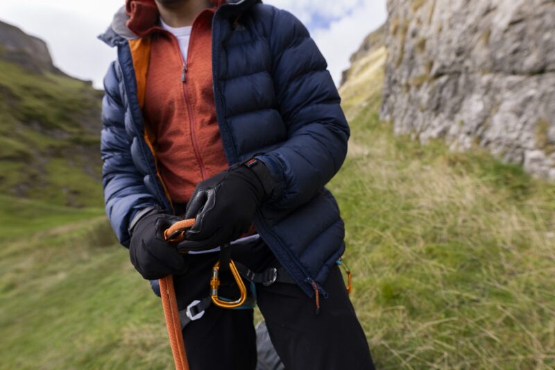 Belay-Gloves-7-3.jpg Person wearing a navy puffer jacket and orange zip-up adjusts an orange climbing rope and gold carabiners with black gloves on a grassy rocky hillside.