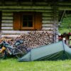 BenReadPhotography_Bikepacking-11-scaled-2.jpg Green one-person tent pitched on grass beside a rustic log cabin with stacked firewood; a fully loaded touring bicycle leans against the cabin and a wooden picnic table with helmets and gear sits nearby, with a grassy forest slope in the background.