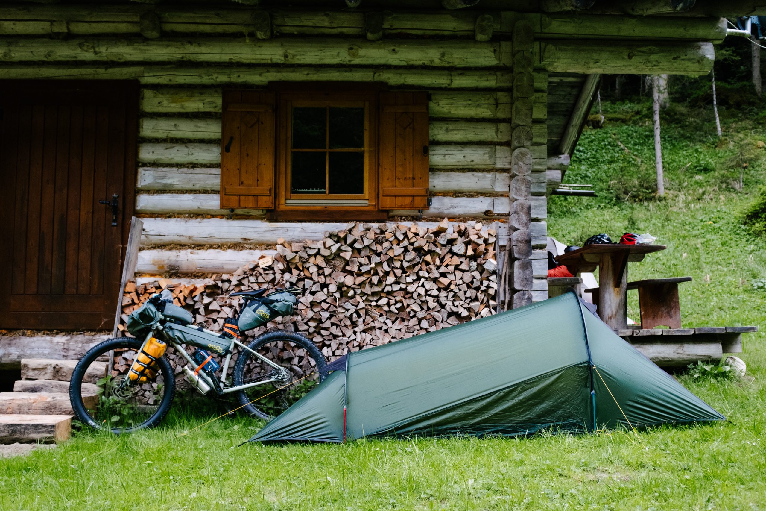 Green one-person tent pitched on grass beside a rustic log cabin with stacked firewood; a fully loaded touring bicycle leans against the cabin and a wooden picnic table with helmets and gear sits nearby, with a grassy forest slope in the background.