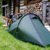 BenReadPhotography_Bikepacking-12-1-scaled-2.jpg Green tunnel tent pitched on grass beside a rustic log cabin and stacked firewood, with a fully loaded touring bicycle leaning against the woodpile and trees in the background