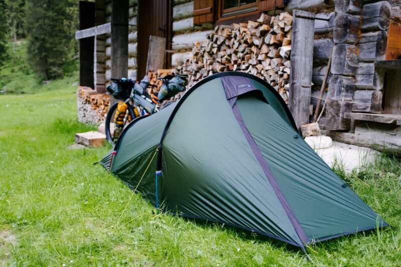 BenReadPhotography_Bikepacking-12-1-scaled-2.jpg Green tunnel tent pitched on grass beside a rustic log cabin and stacked firewood, with a fully loaded touring bicycle leaning against the woodpile and trees in the background