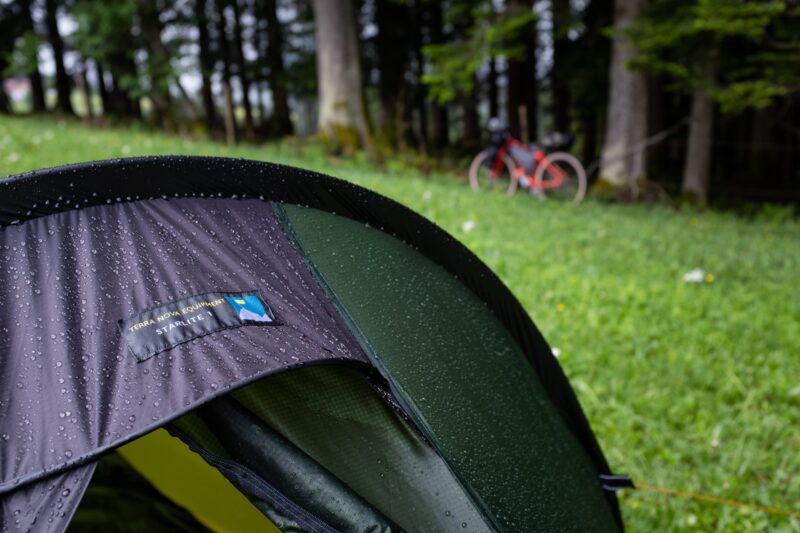 BenReadPhotography_Bikepacking-6-scaled-2.jpg Close-up of a rain-speckled green and grey Terra Nova Equipment Starlite tent fly pitched on wet grass, with a blurred red bicycle and trees in the background.