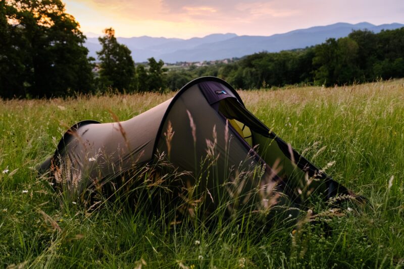 BenReadPhotography_Bikepacking-89-scaled-2.jpg Olive-green tunnel tent pitched in a tall grassy meadow with wildflowers, surrounded by trees and distant blue mountains beneath a pink-orange sunset sky.