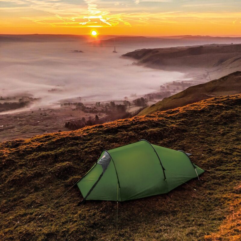 Green tunnel tent pitched on a grassy ridge at sunrise, overlooking a fog-filled valley with low mist and a distant chimney-like tower beneath an orange sky.