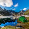 Green camping tent pitched on a grassy, rocky shore beside a clear alpine lake reflecting jagged, snow-dusted mountains and sweeping clouds under a vivid blue sky.