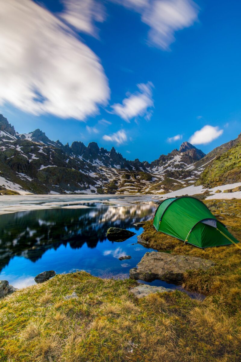 Green camping tent pitched on a grassy, rocky shore beside a clear alpine lake reflecting jagged, snow-dusted mountains and sweeping clouds under a vivid blue sky.