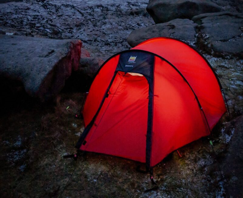 Red dome tent glowing from an interior light, pitched on frosty ground among large weathered rocks at dusk.