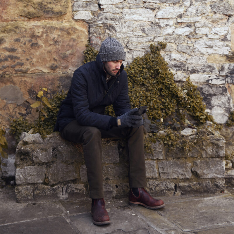 Bora-and-Baslow-1080x1080-1-1.jpg A man in a grey knit hat, navy quilted jacket and gloves sits on a low stone wall with ivy, looking at a smartphone while wearing brown trousers and reddish-brown boots against a weathered stone wall.