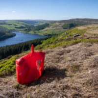 Bright red bivvy bag with a small clear window propped on a dry hillside overlooking a blue reservoir, dense pine woodland and patchwork green hills beneath a clear sky.