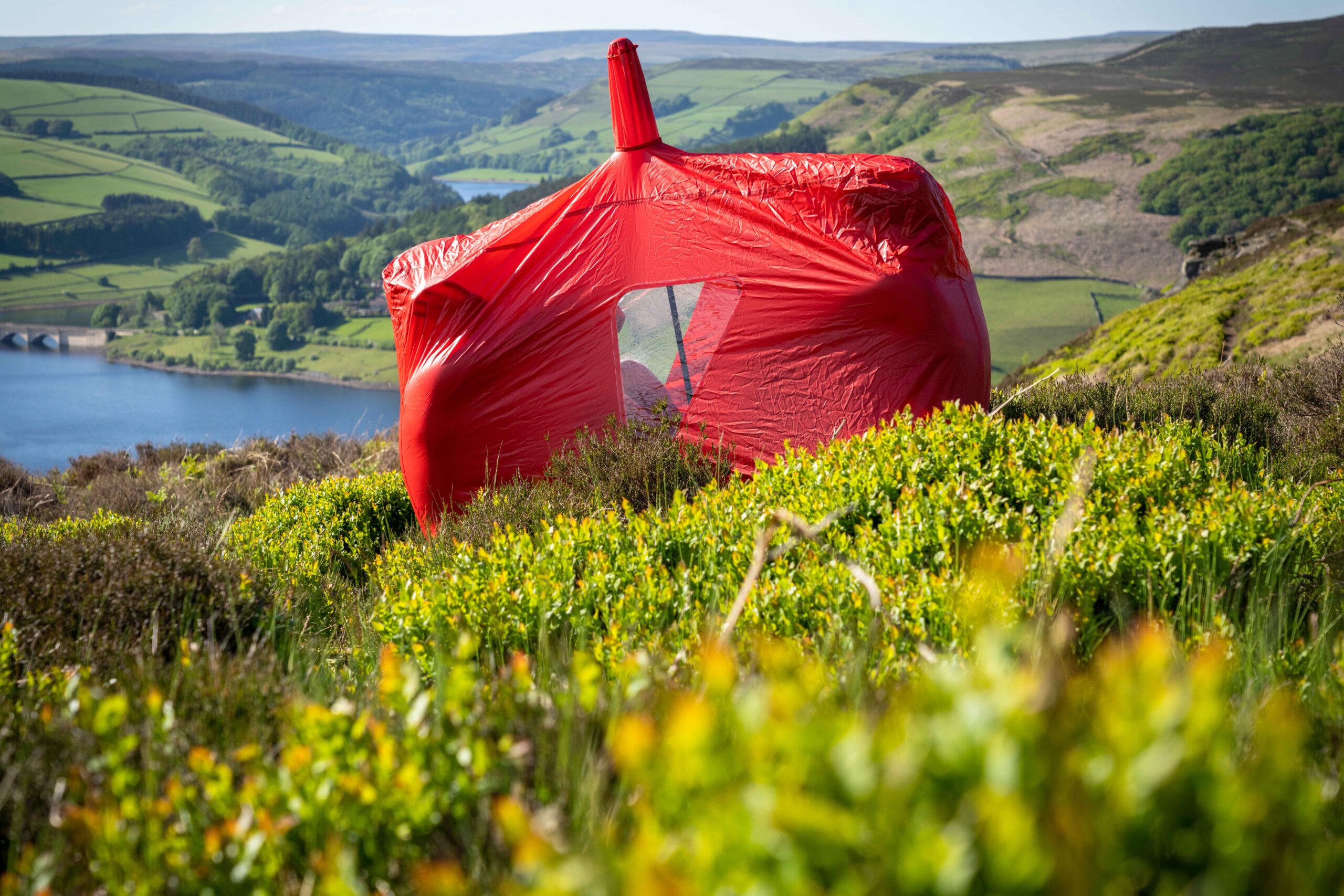 Terra Nova Shoot A bright red tent wrapped in a taut protective cover sits among green heather on a sunlit moorland slope, overlooking a blue reservoir and patchwork rolling hills.