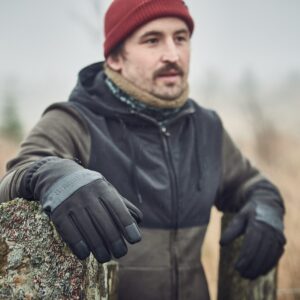 A man wearing a red beanie, scarf and dark jacket leans on a lichen-covered stone gatepost in a misty field, his black-gloved hand prominent in the foreground.