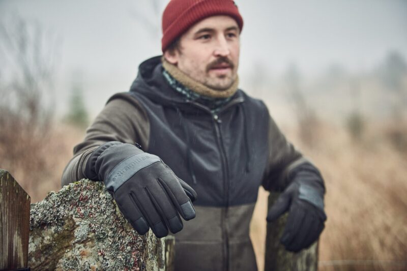 Bullet-Glove-22BLTG-2-scaled-4.jpg A man wearing a red beanie, scarf and dark jacket leans on a lichen-covered stone gatepost in a misty field, his black-gloved hand prominent in the foreground.