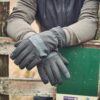 Bullet-Glove-22BLTG-scaled-4.jpg A pair of dark grey insulated gloves on crossed hands resting over a weathered wooden fence next to a green sign, the wearer's grey jacket sleeves visible.
