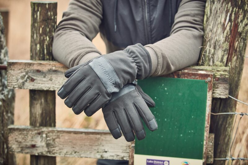 Bullet-Glove-22BLTG-scaled-4.jpg A pair of dark grey insulated gloves on crossed hands resting over a weathered wooden fence next to a green sign, the wearer's grey jacket sleeves visible.