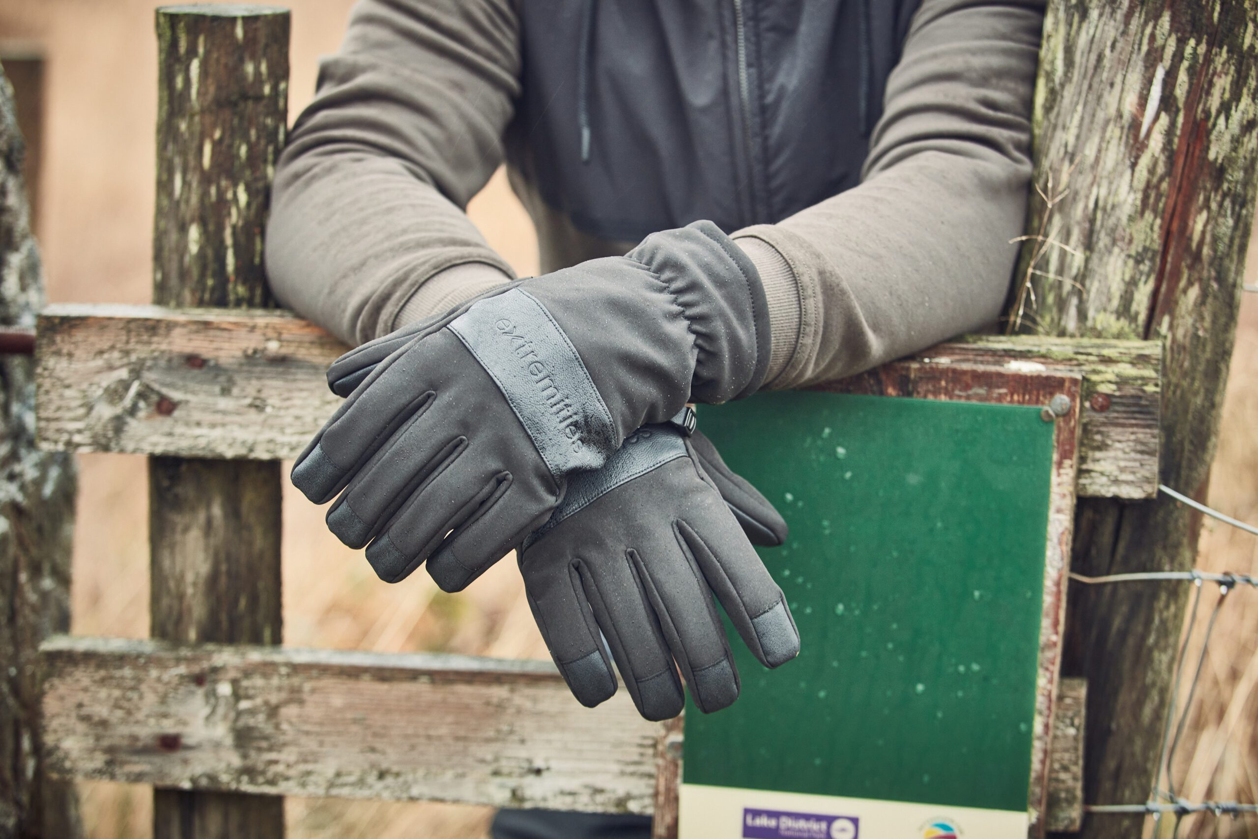 A pair of dark grey insulated gloves on crossed hands resting over a weathered wooden fence next to a green sign, the wearer's grey jacket sleeves visible.