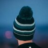 Capella-Beanie-Dark-1500-px-1.jpg Rear view of a person wearing a knitted teal and dark green striped beanie with a large pom-pom against a soft-focus blue background.