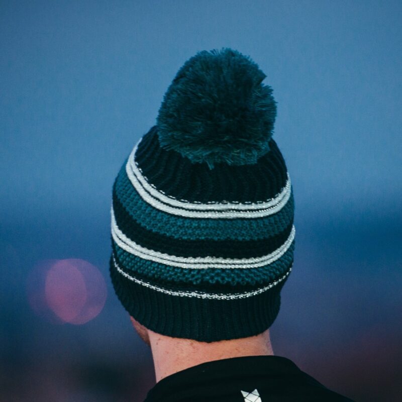 Capella-Beanie-Dark-1500-px-1.jpg Rear view of a person wearing a knitted teal and dark green striped beanie with a large pom-pom against a soft-focus blue background.
