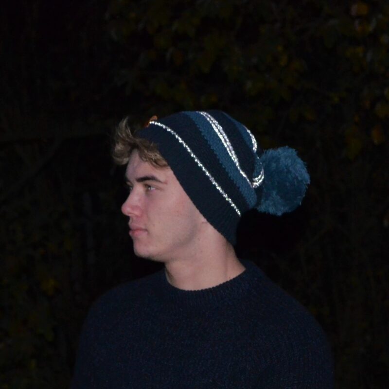 Capella-Beanie-in-the-dark-cropped-1500px-1.jpg A young man in profile wearing a navy and teal striped knit beanie with a large pompom and a dark sweater, set against a dim, leafy background.