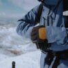 Capitol-Peak-1-scaled-4.jpg Person in a blue winter jacket and harness wearing black-and-yellow insulated gloves clasping their hands while standing on snowy mountain slopes with ski-pole handles visible.