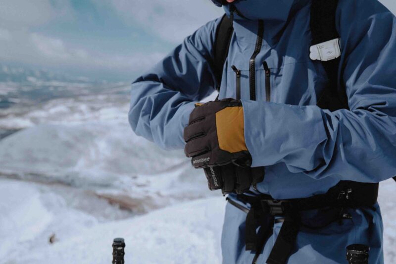 Person in a blue winter jacket and harness wearing black-and-yellow insulated gloves clasping their hands while standing on snowy mountain slopes with ski-pole handles visible.
