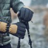 Capitol-Peak-22CAP-Lifestyle-4-scaled-4.jpg A person wearing a green quilted jacket grips trekking poles with black-and-tan insulated mittens dotted with raindrops on a wet, blurred outdoor path.