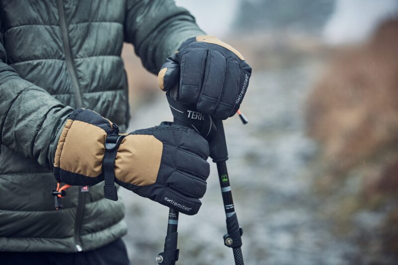 A person wearing a green quilted jacket grips trekking poles with black-and-tan insulated mittens dotted with raindrops on a wet, blurred outdoor path.