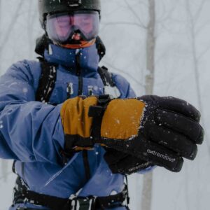 Close-up of a person in a blue ski jacket, helmet and pink goggles adjusting orange-and-black insulated gloves labeled "extremities" as snow falls in a bare winter forest