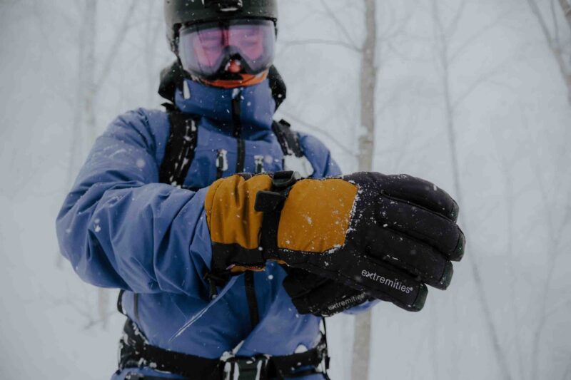 Close-up of a person in a blue ski jacket, helmet and pink goggles adjusting orange-and-black insulated gloves labeled "extremities" as snow falls in a bare winter forest