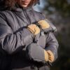 Close-up of a person wearing a dark puffer jacket and tan-and-grey insulated mittens clipped together, arms crossed against an outdoor background.
