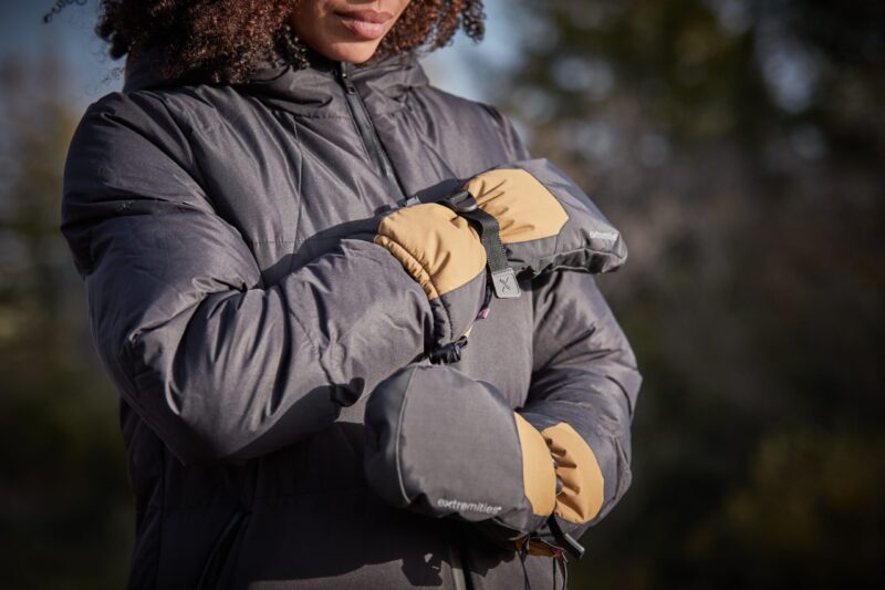 Close-up of a person wearing a dark puffer jacket and tan-and-grey insulated mittens clipped together, arms crossed against an outdoor background.