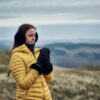 Chamonix-Glove-22CHG-scaled-4.jpg Woman wearing a yellow puffer jacket, black earmuffs and gloves standing on windswept grassy hills, clasping her hands as misty blue‑grey ridges and an overcast sky recede behind her.