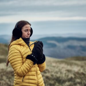 Woman wearing a yellow puffer jacket, black earmuffs and gloves standing on windswept grassy hills, clasping her hands as misty blue‑grey ridges and an overcast sky recede behind her.