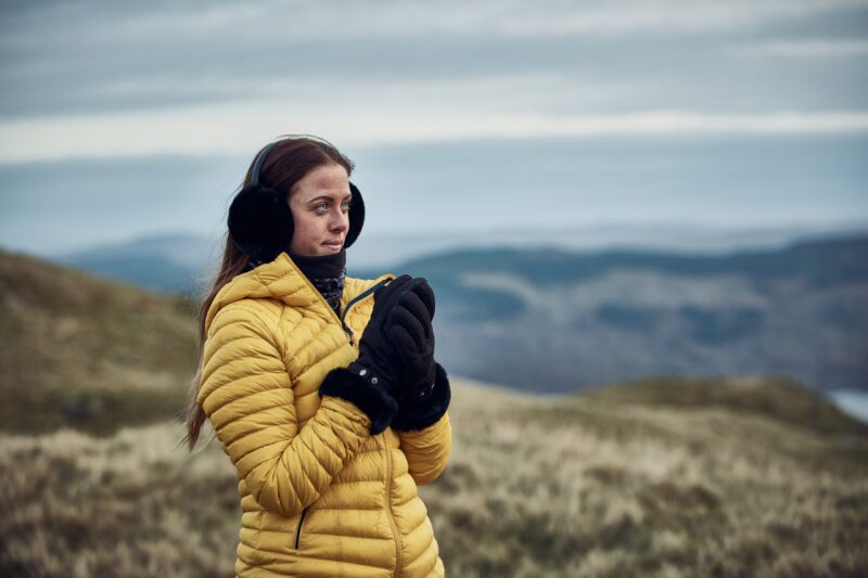 Chamonix-Glove-22CHG-scaled-4.jpg Woman wearing a yellow puffer jacket, black earmuffs and gloves standing on windswept grassy hills, clasping her hands as misty blue‑grey ridges and an overcast sky recede behind her.