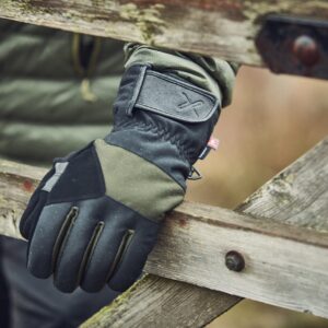 Close-up of a black and olive-green outdoor glove with a Velcro wrist strap and tiny red tag resting on a weathered wooden gate, rain droplets visible on the glove and jacket sleeve behind it.