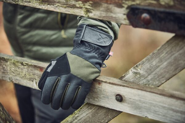 Commando-Glove-22CMG-2-scaled-4.jpg Close-up of a black and olive-green outdoor glove with a Velcro wrist strap and tiny red tag resting on a weathered wooden gate, rain droplets visible on the glove and jacket sleeve behind it.