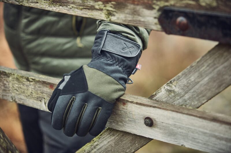 Close-up of a black and olive-green outdoor glove with a Velcro wrist strap and tiny red tag resting on a weathered wooden gate, rain droplets visible on the glove and jacket sleeve behind it.