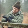Commando-Glove-22CMG-scaled-4.jpg Young man wearing an olive beanie, colourful neck gaiter and green quilted jacket adjusts black winter gloves while leaning on a moss-covered wooden gate in a misty woodland.