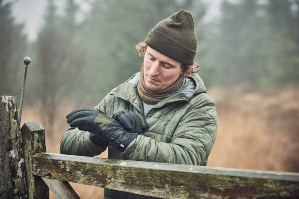 Commando-Glove-22CMG-scaled-4.jpg Young man wearing an olive beanie, colourful neck gaiter and green quilted jacket adjusts black winter gloves while leaning on a moss-covered wooden gate in a misty woodland.