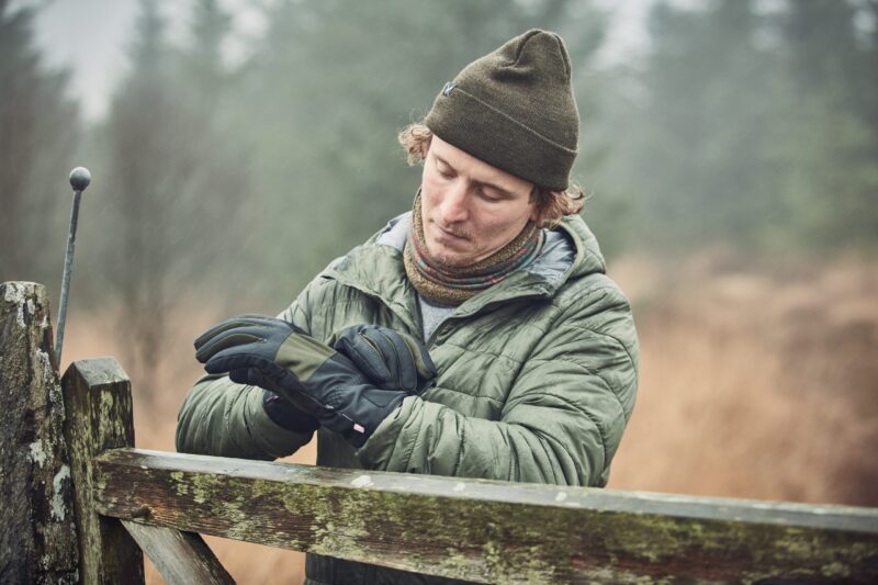 Young man wearing an olive beanie, colourful neck gaiter and green quilted jacket adjusts black winter gloves while leaning on a moss-covered wooden gate in a misty woodland.