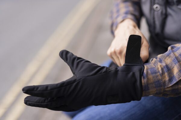 Commuter-Gloves-1-3.jpg Close-up of a person fastening the Velcro strap of a black glove over their wrist while wearing a blue-and-brown checked shirt, with blurred road lines in the background.